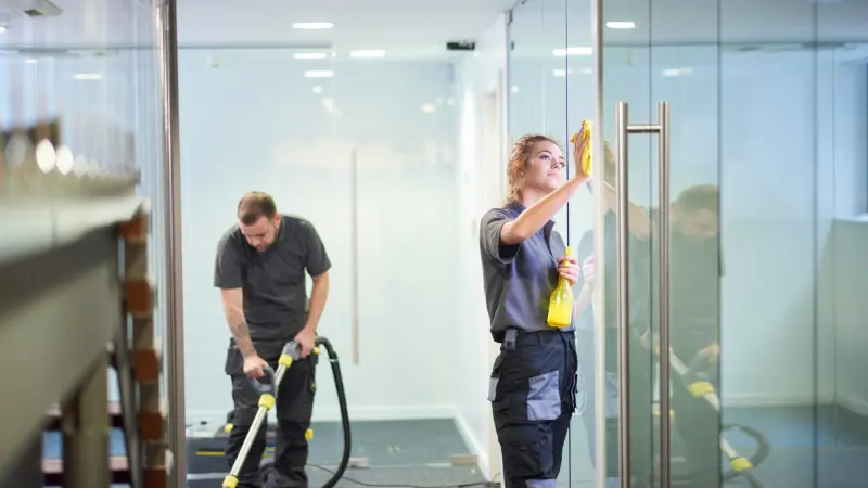 Two professional BJR cleaners in grey uniforms working in a modern Perth office — one wiping a glass partition, the other vacuuming carpet.